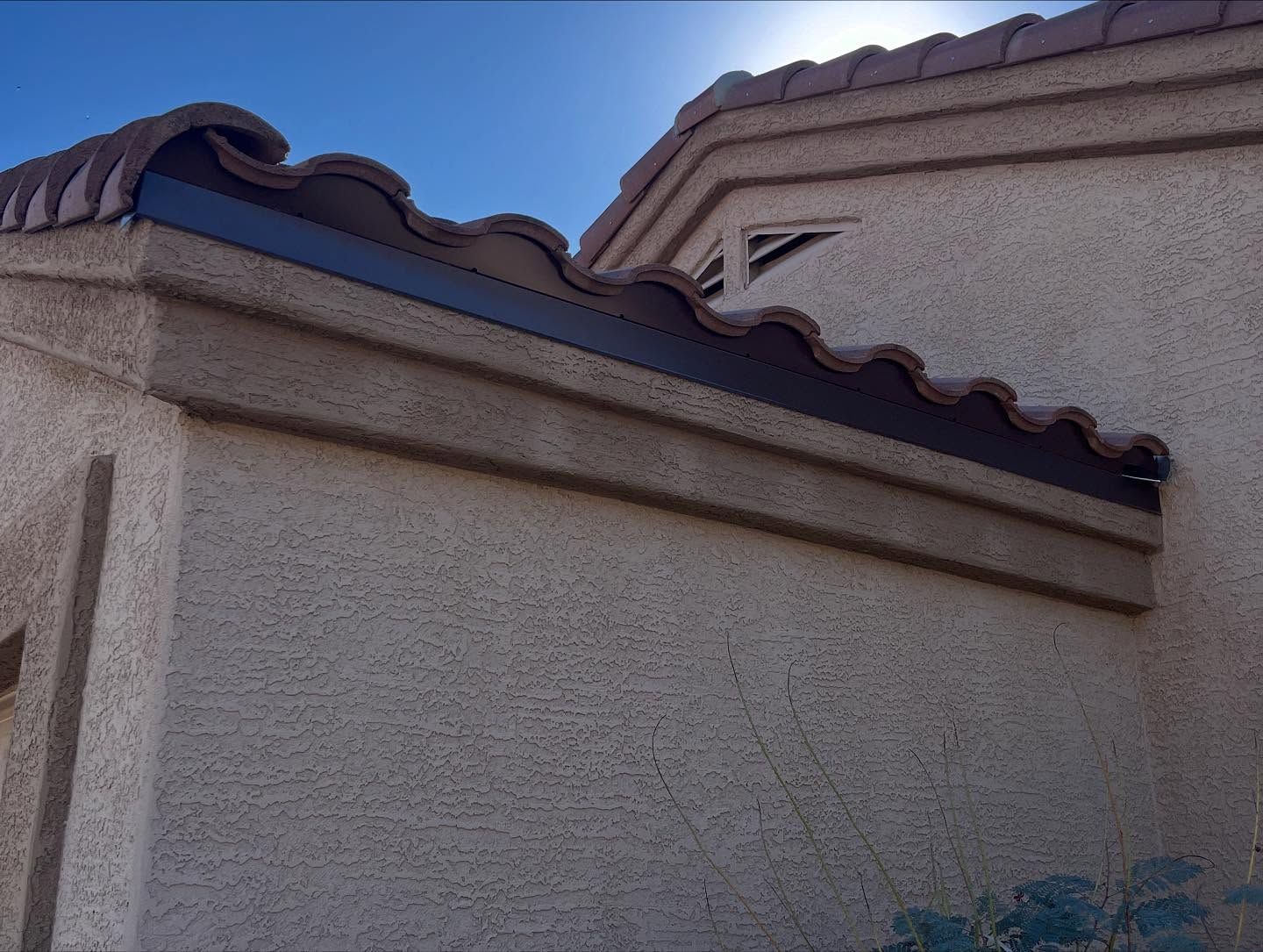Tan stucco wall with brown trim and Spanish tile roof under a bright sky.
