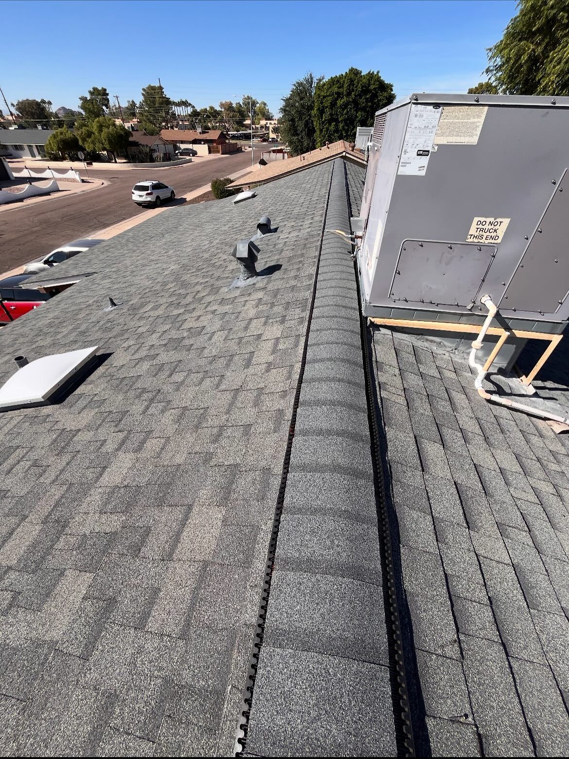 Asphalt shingle roof with vents, a central ridge cap, and an AC unit, on a sunny day.