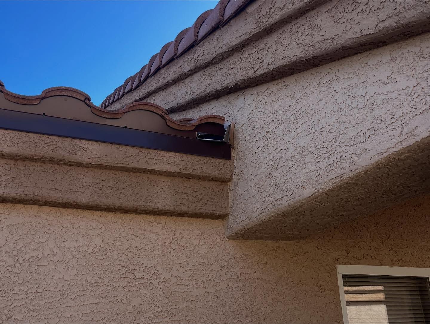 Close-up of stucco exterior with brown roof tiles and trim against a clear blue sky.