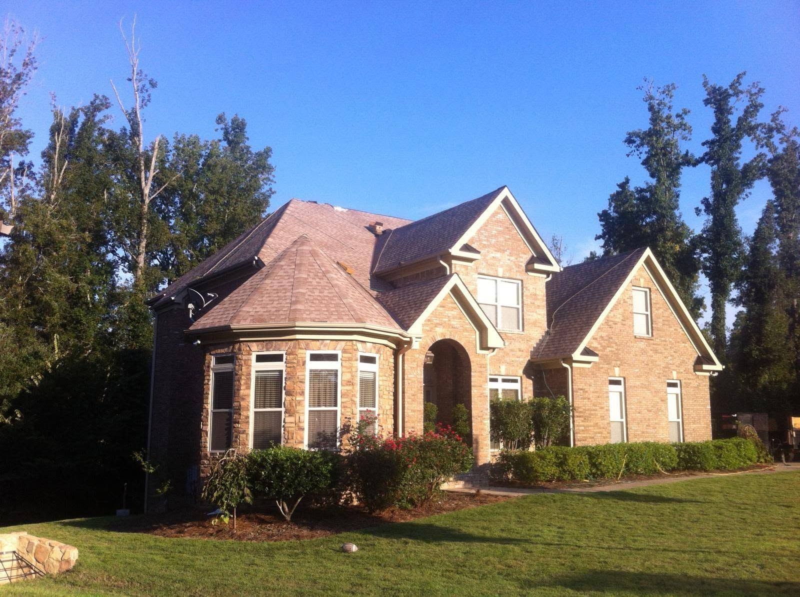 Brick house with a red roof and large windows on a sunny lawn, surrounded by trees.