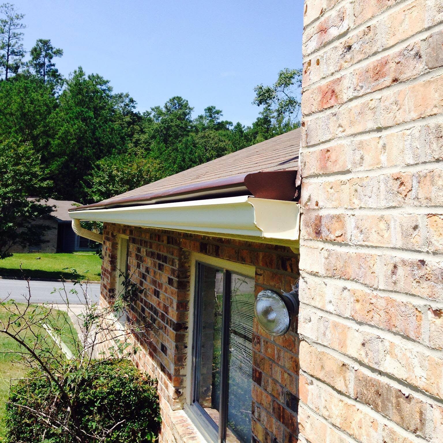 Brick house exterior with tan roof and white trim, viewed beside a driveway and trees.
