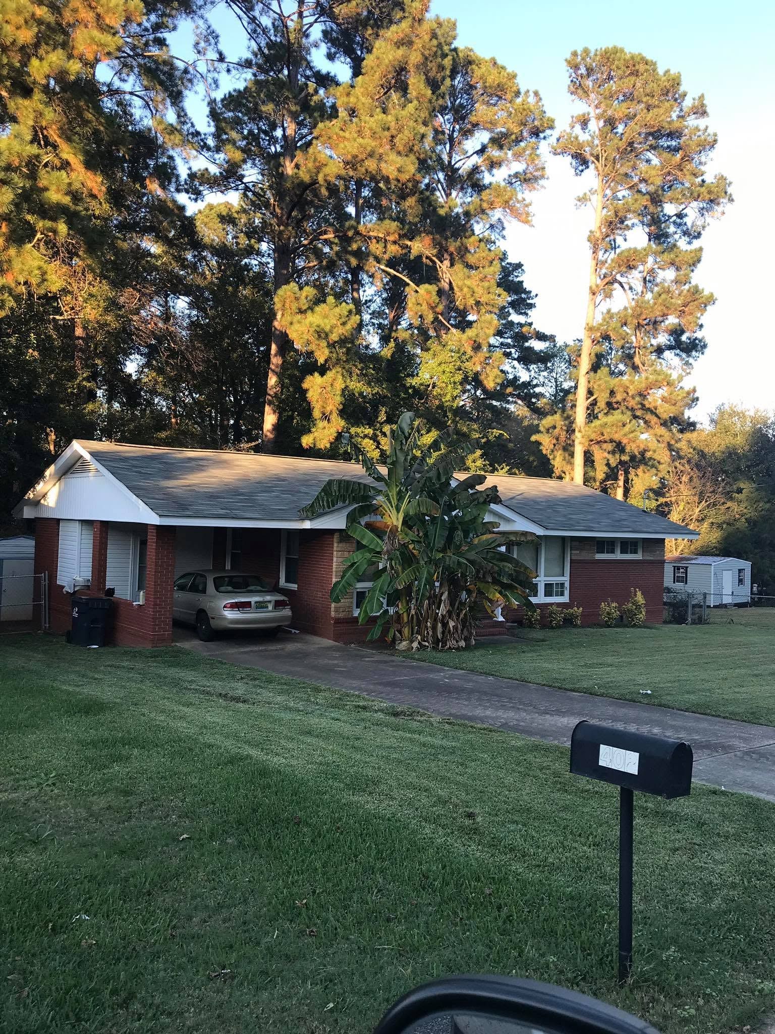 Brick house with carport, lawn, and mailbox at sunset, backed by tall trees.