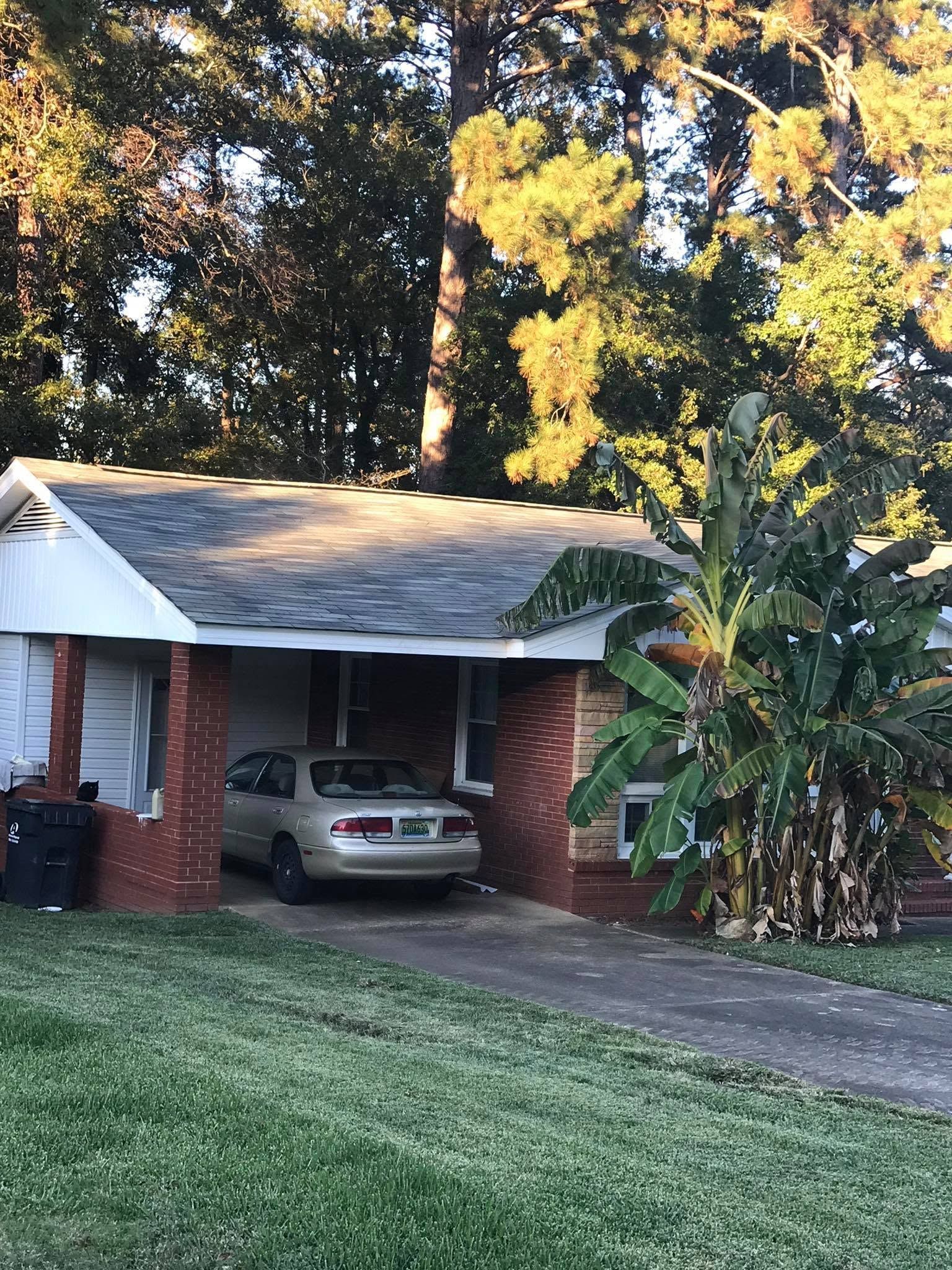 Brick house with a car in the driveway, surrounded by tall trees and greenery.