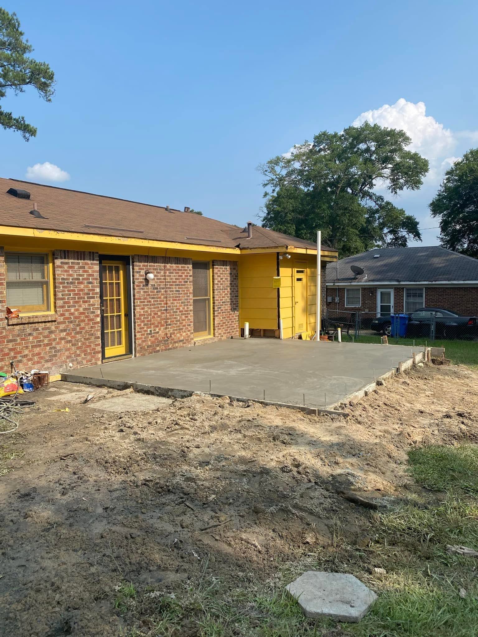 New concrete patio beside a brick house with yellow trim, viewed from a dirt yard.