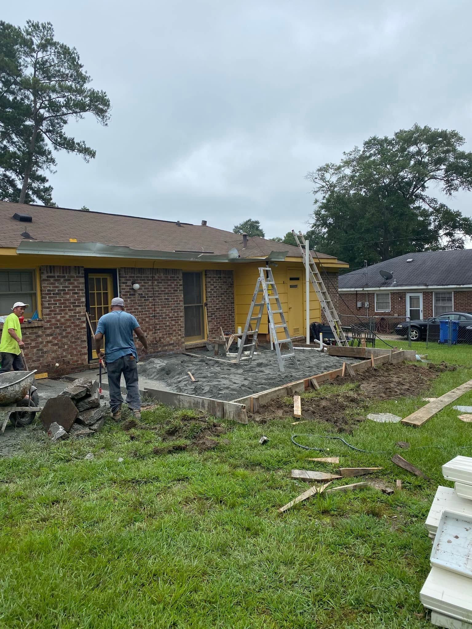Workers renovating a brick house exterior with ladders, gravel, and a yellow door on a cloudy day