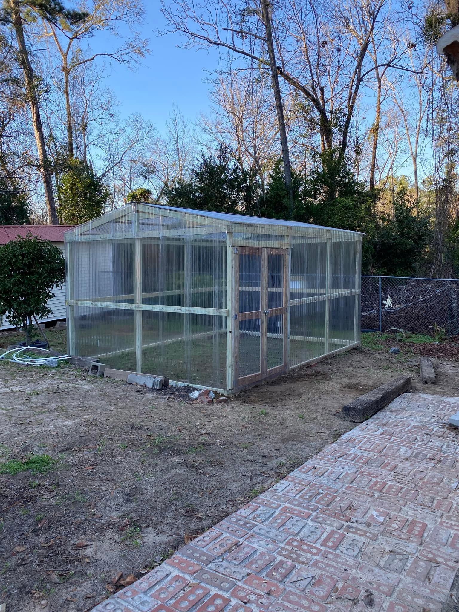 Small glass greenhouse in a backyard beside a brick patio, surrounded by trees and gravel.