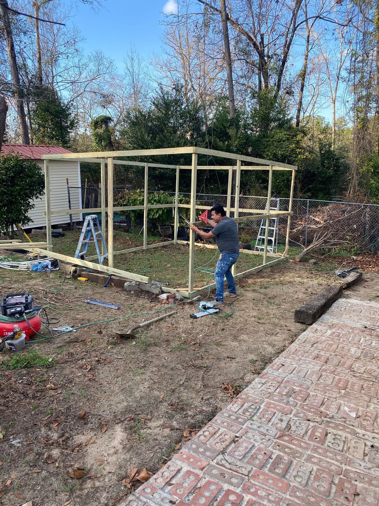 Person building a wooden shed frame in a backyard next to a brick patio.