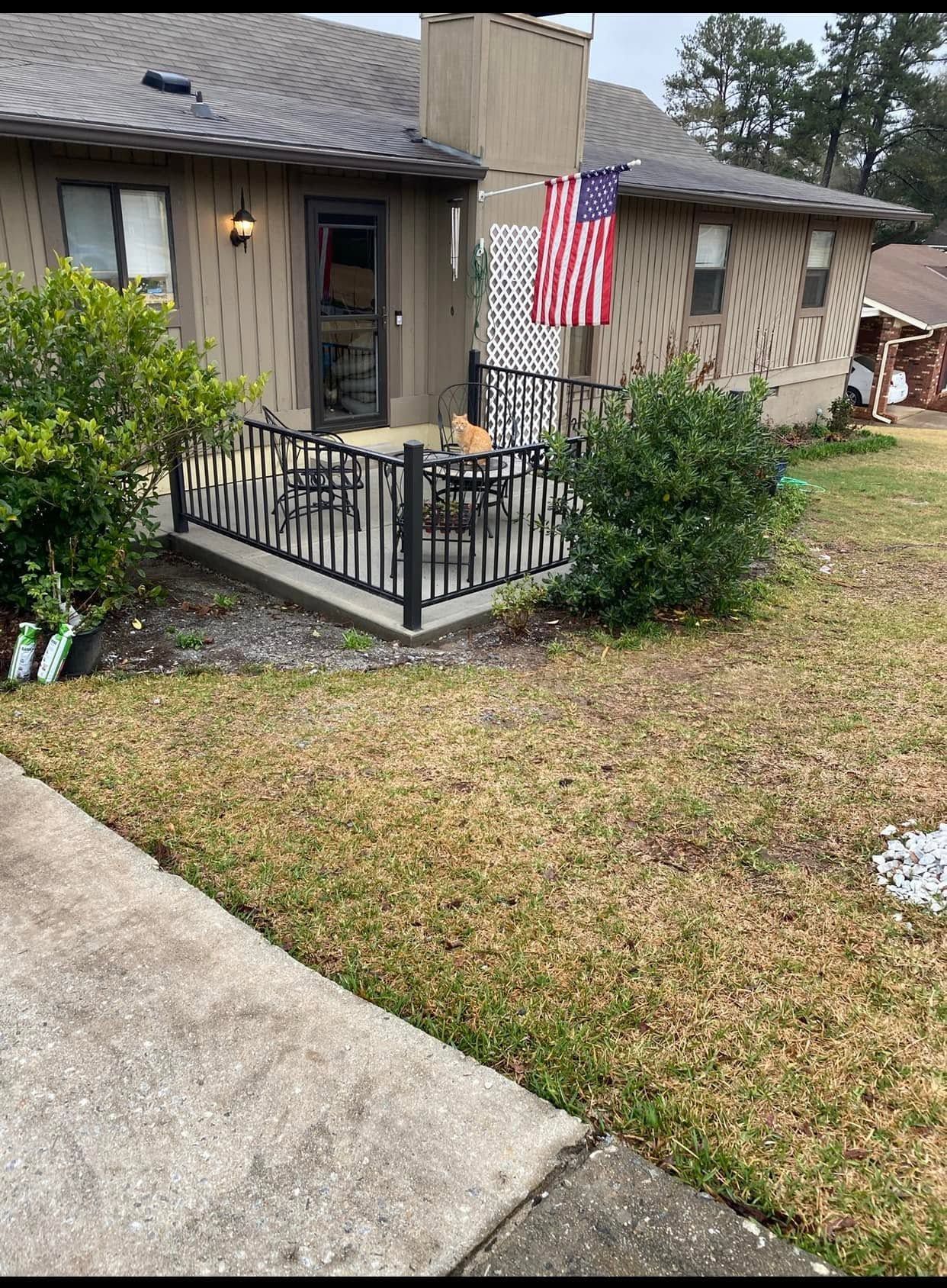 Front porch with black railing, American flag, and shrubs beside a grassy yard and sidewalk.