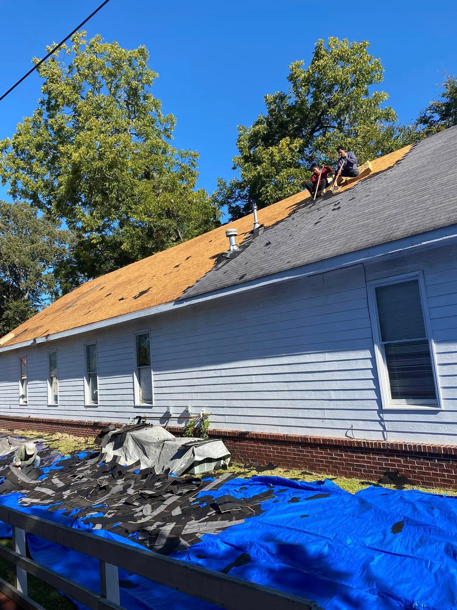 White house with gray roof being reroofed, orange underlayment exposed, blue tarp and debris in front.