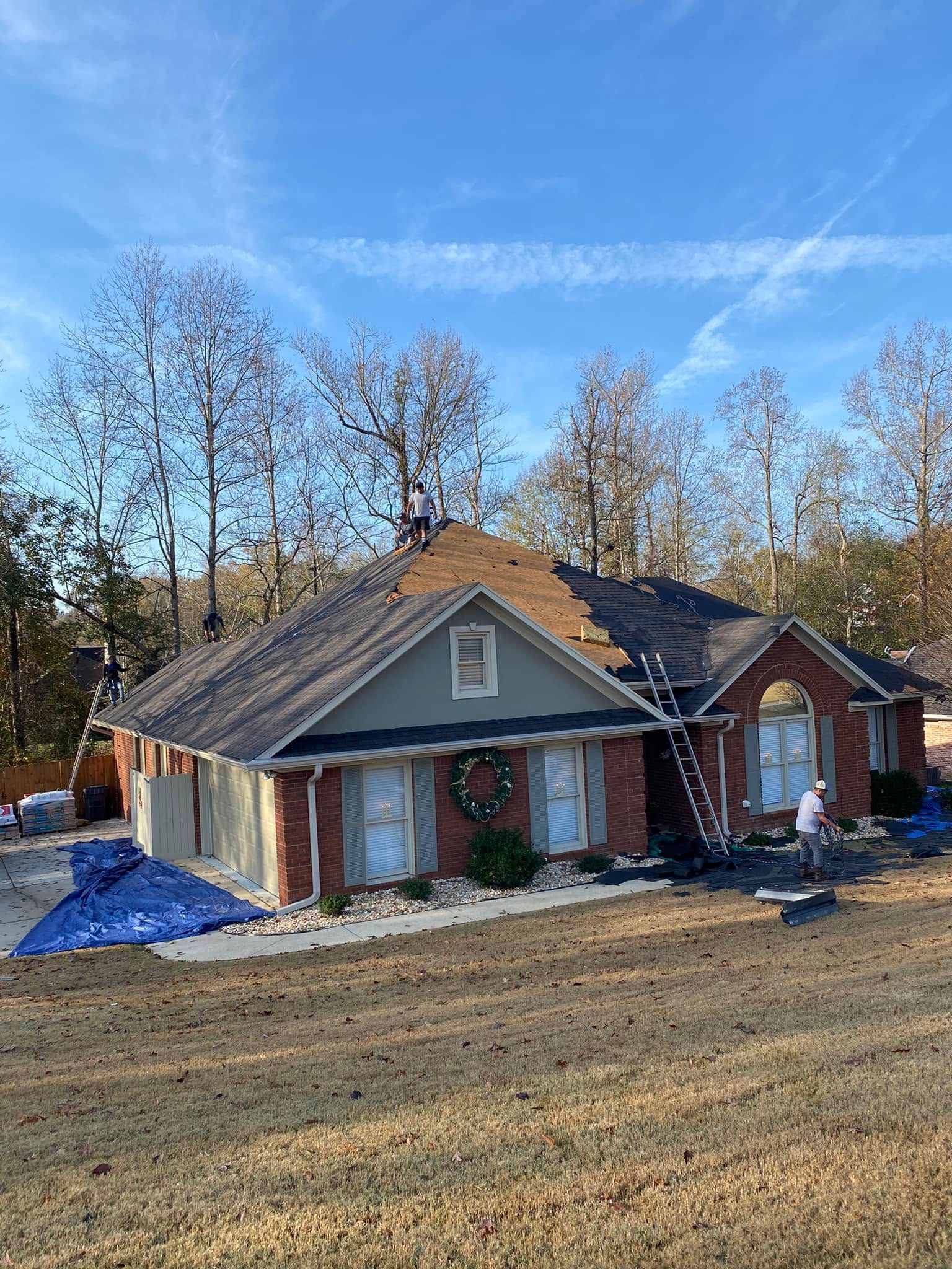 Damaged single-story brick house with exposed roof under a clear blue sky, surrounded by bare trees.
