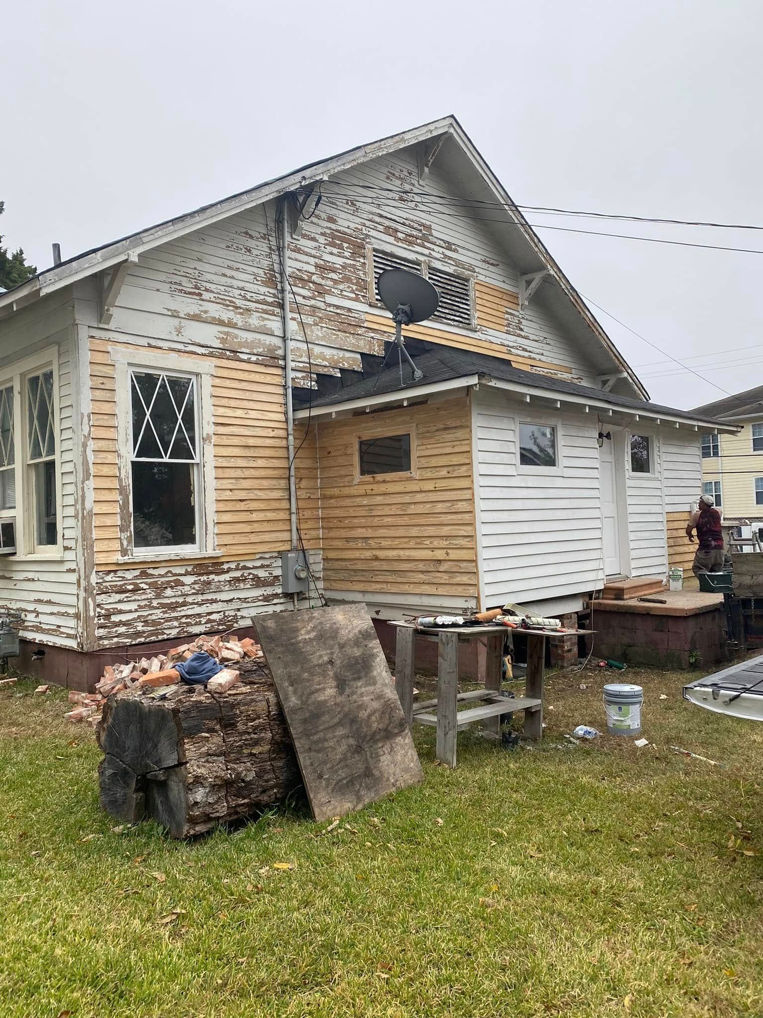 Damaged house exterior with missing siding, boarded windows, and debris in the yard