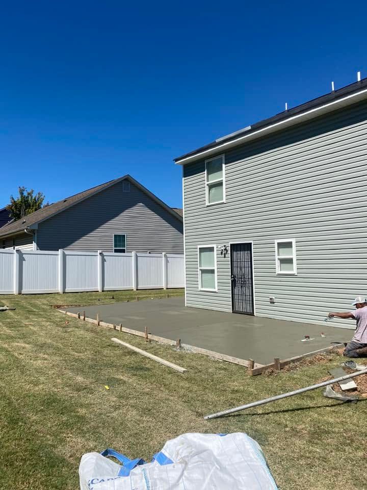 Backyard with gray house, white fence, grass lawn, and a person working near a paved patio