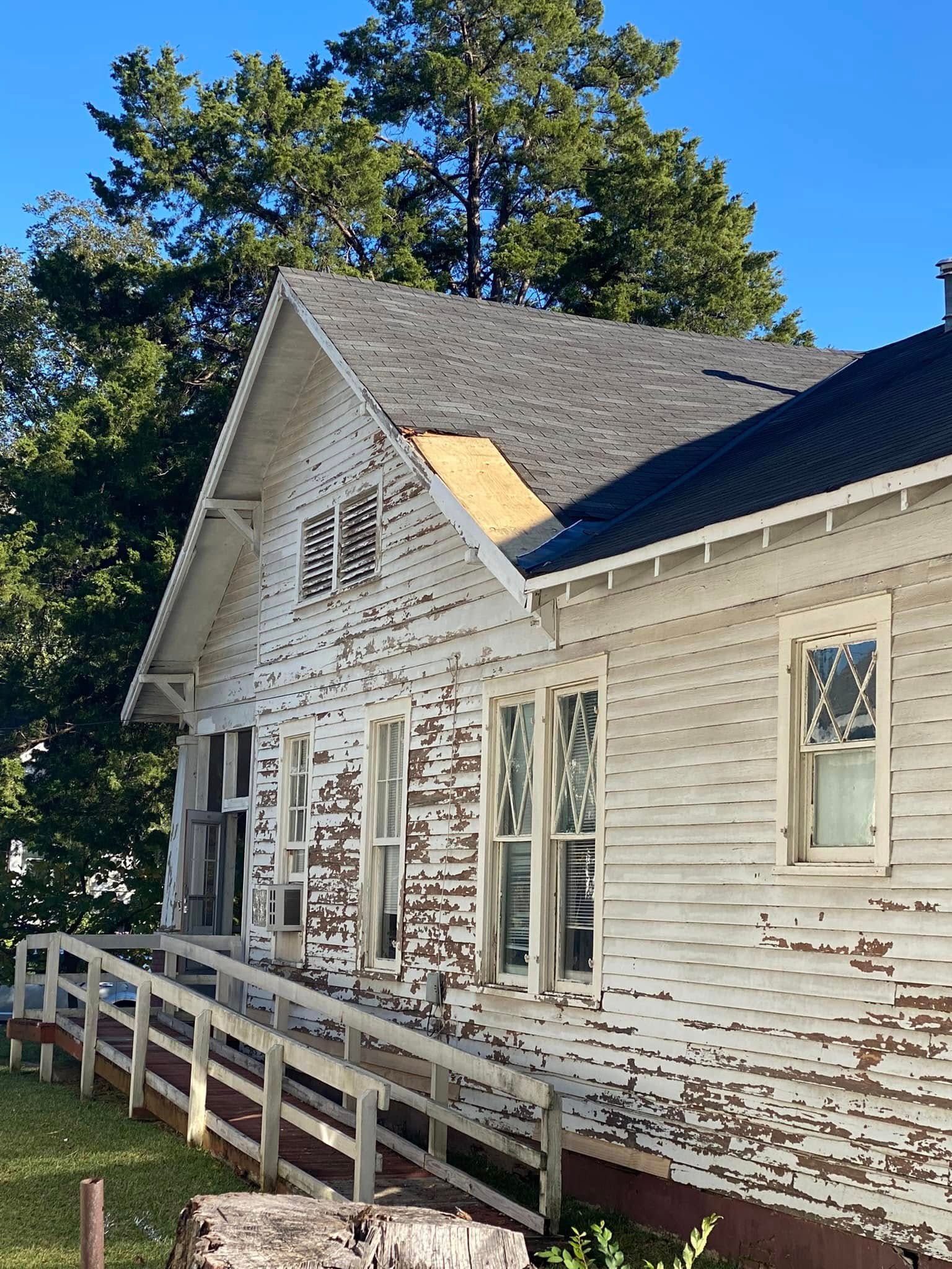 Weathered white house with porch ramp, peeling paint, and tall trees in the background