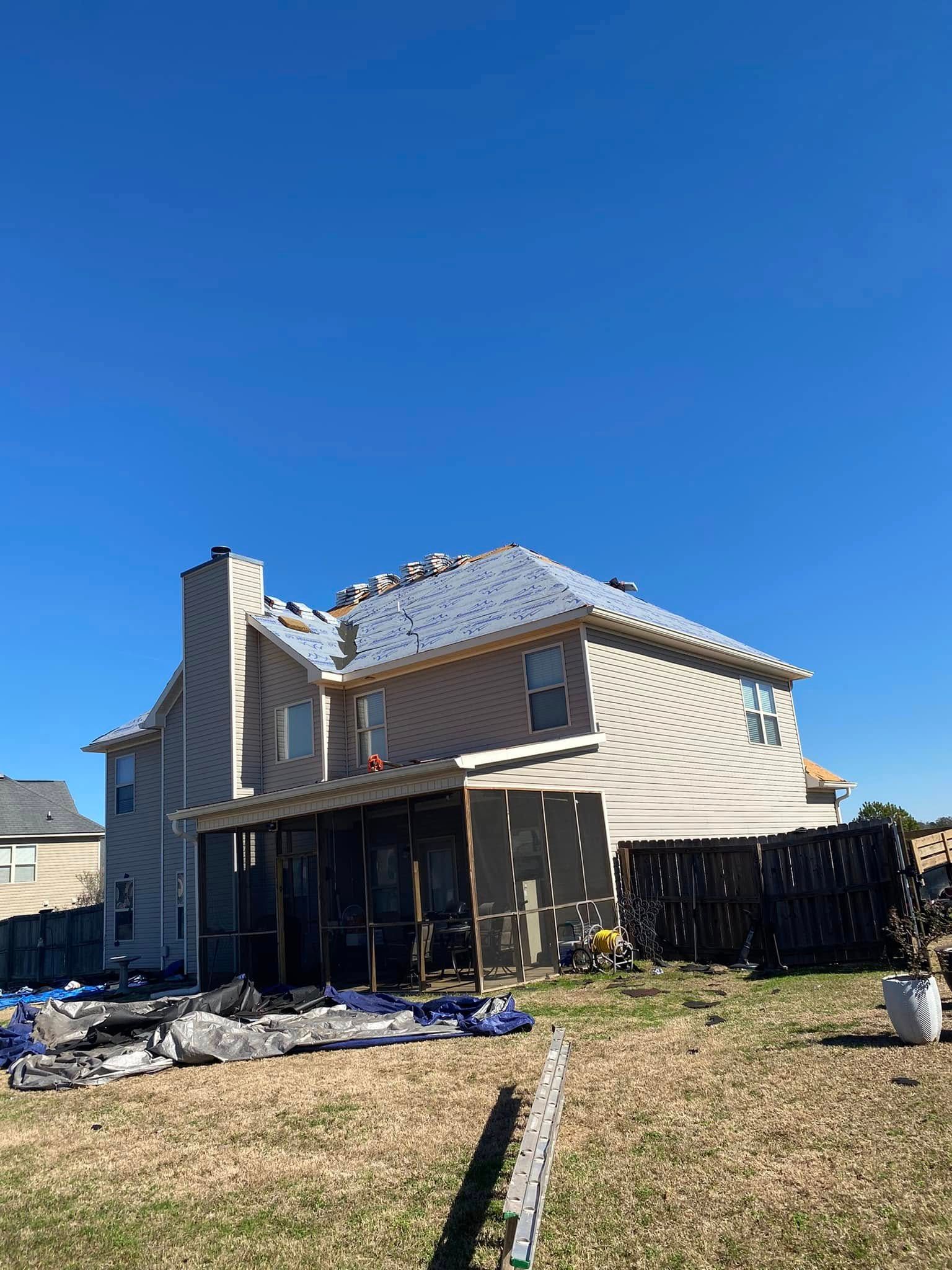 Damaged two-story house with roof tiles missing and debris in the yard under a clear blue sky