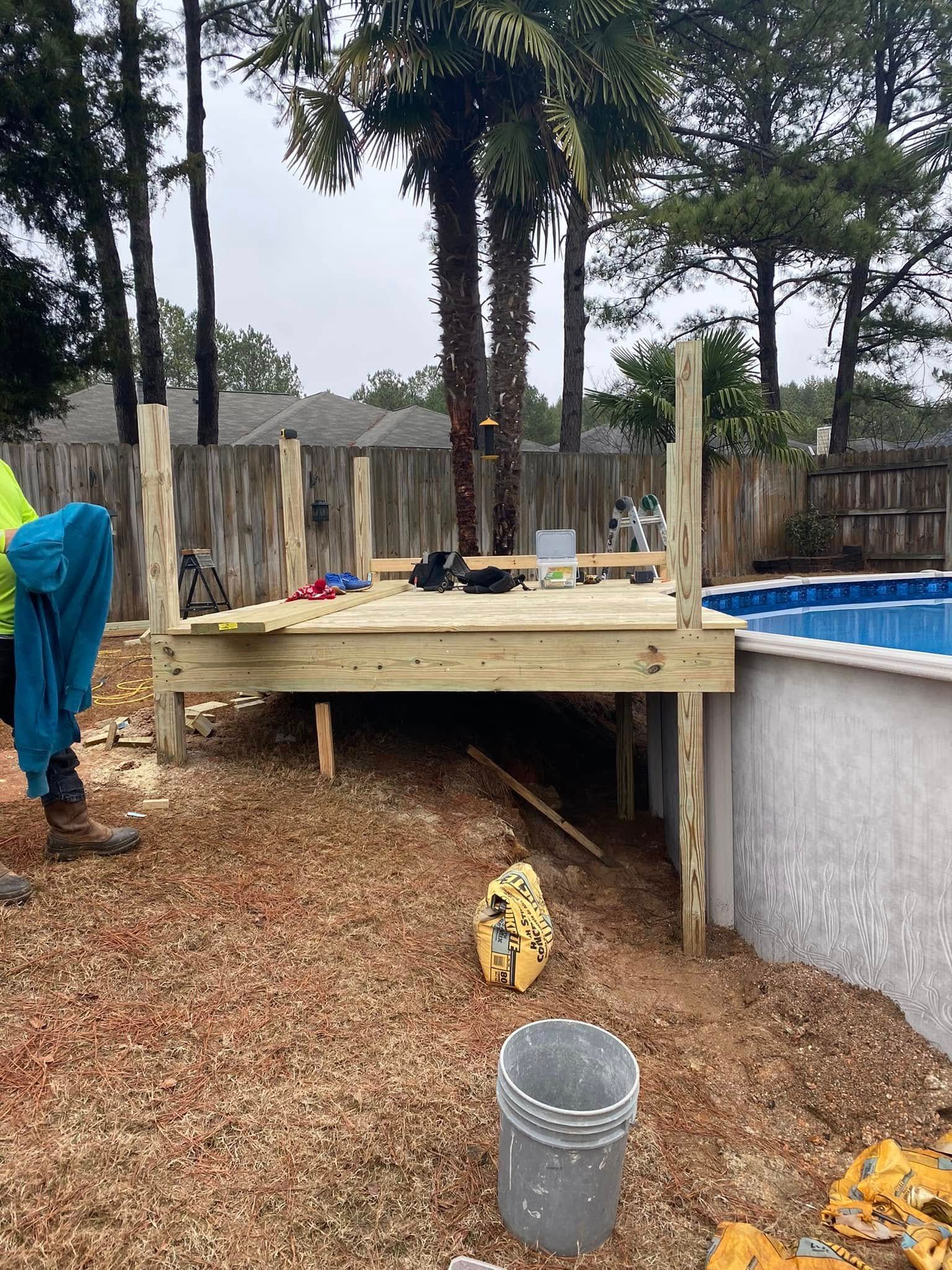 Backyard raised deck under construction beside an above-ground pool, with tools and a bucket on the ground.