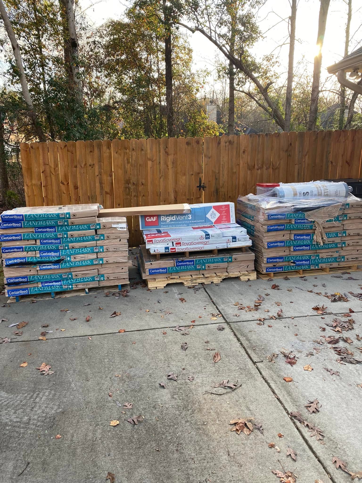 Stacks of packaged lumber or paving stones on pallets beside a wooden fence in a driveway