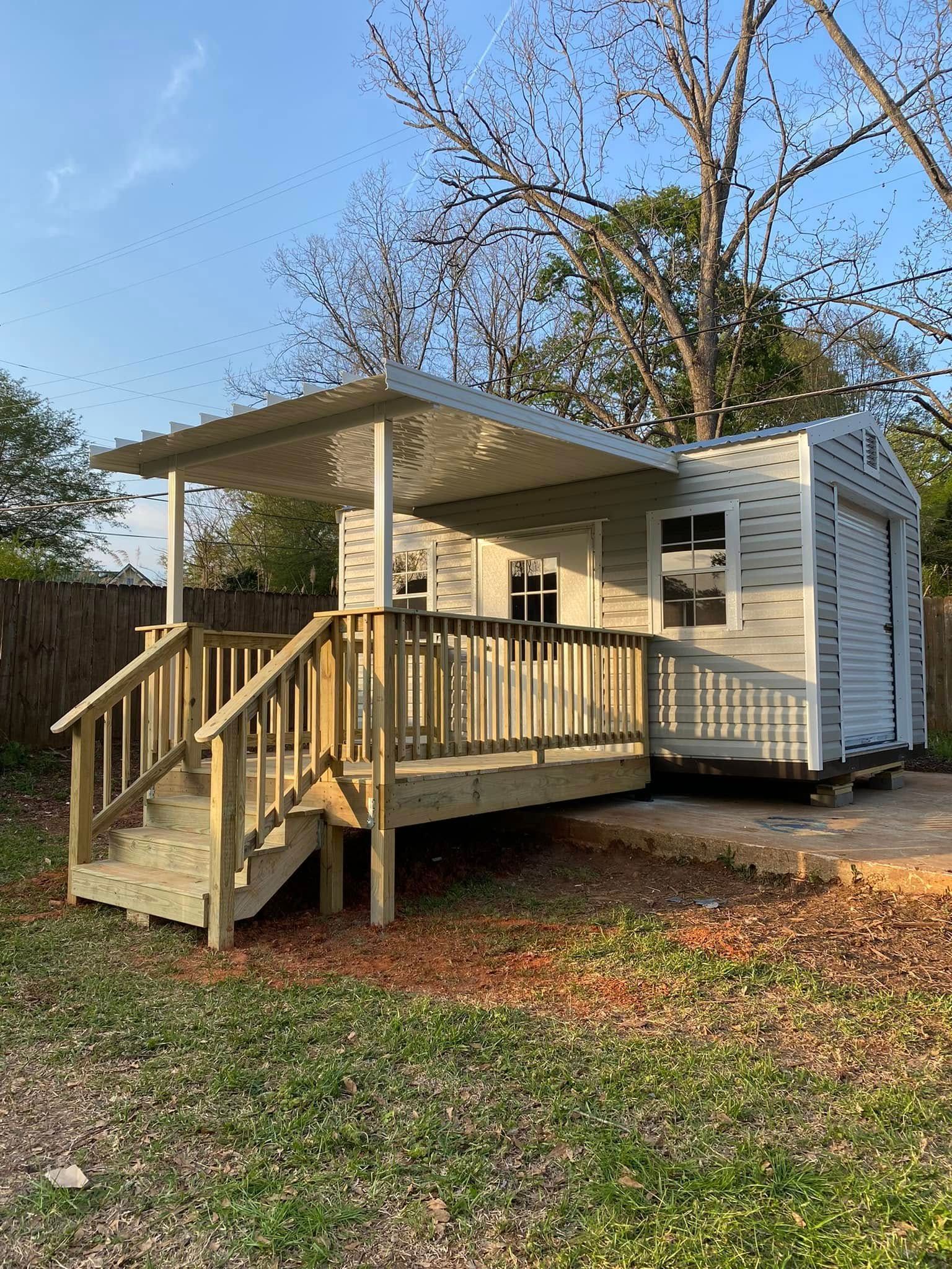 Small beige house with a front porch and wooden ramp, surrounded by trees under a blue sky