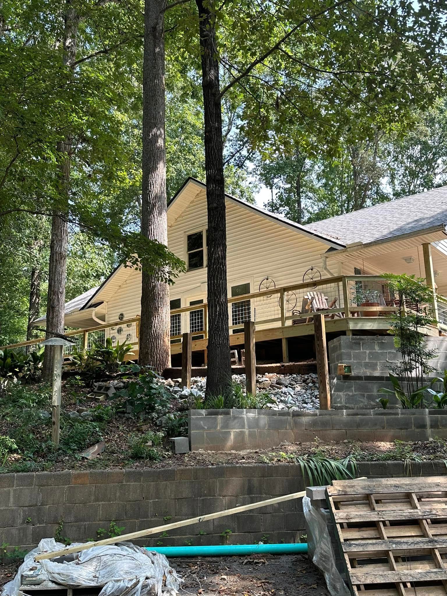 Wooded hillside with a tan house on stilts and a stone retaining wall above a stream.