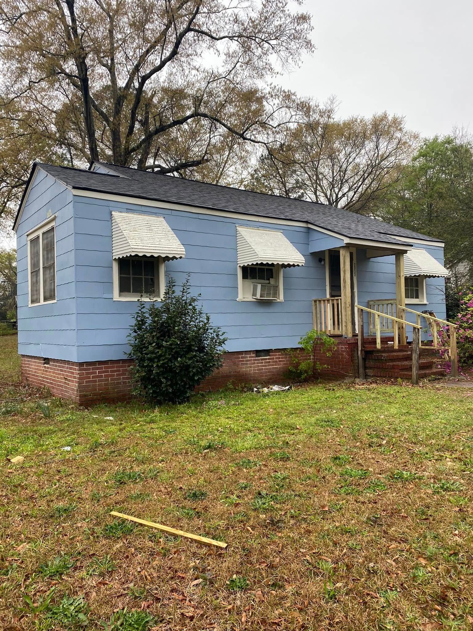Small blue house with white awnings and brick foundation, seen in a leafy yard on an overcast day