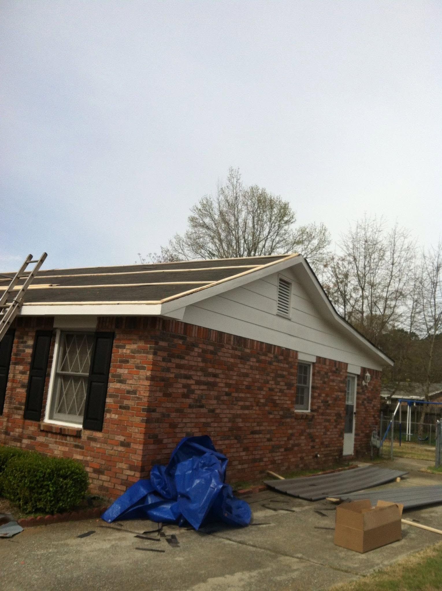 Brick house with gray roof, blue tarp on the lawn, and a ladder near the left side.