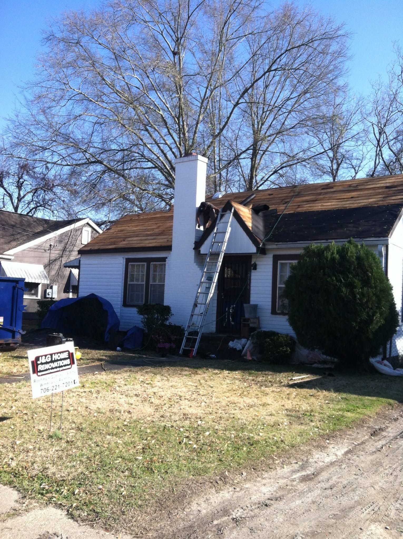 Small white house with a tall chimney, brown roof, bare tree, and yard sign in front