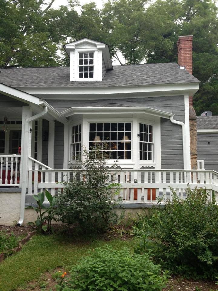 Gray house with white trim, porch, dormer window, and red brick chimney amid green trees and shrubs