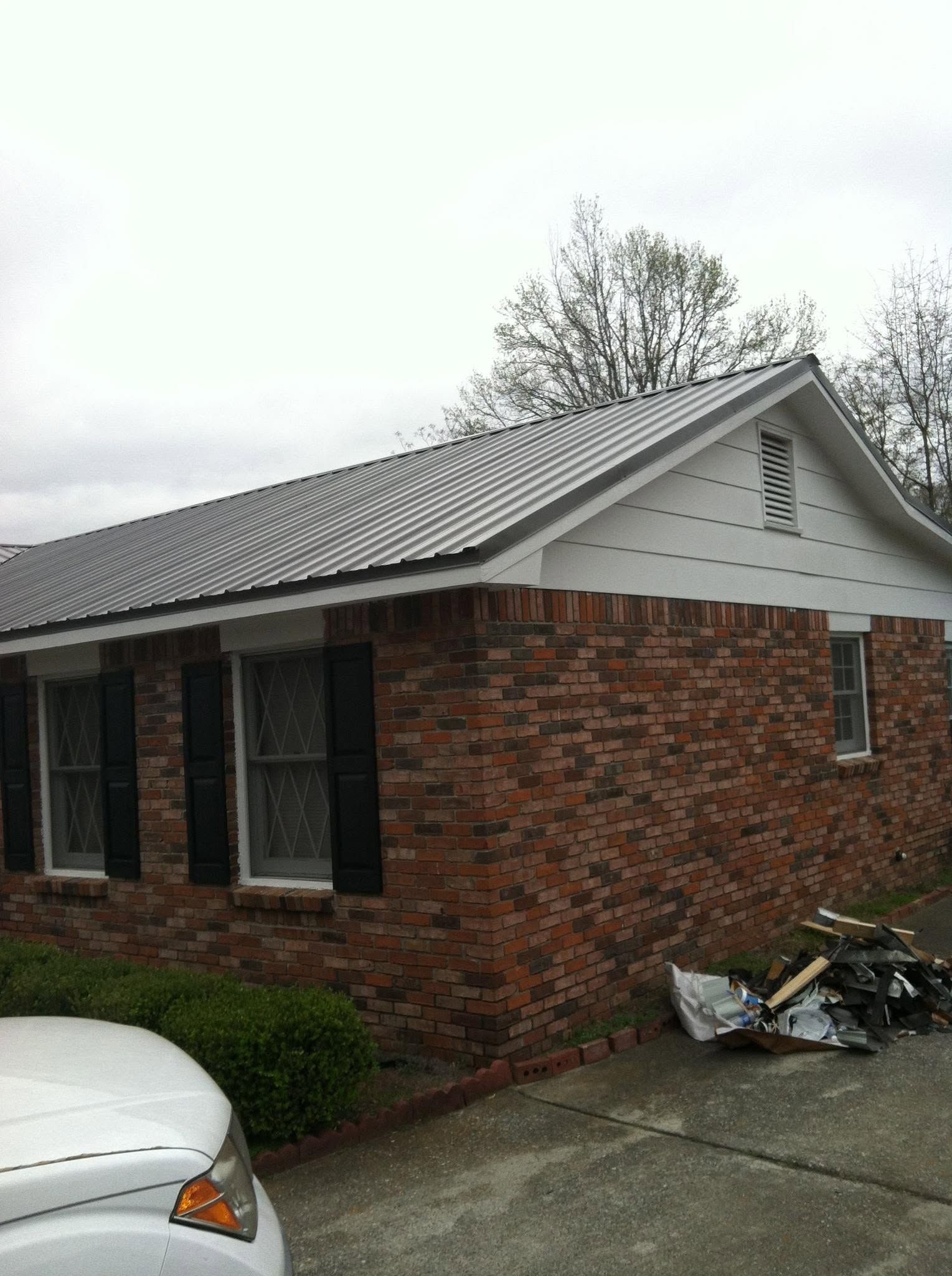 Brick house with gray metal roof, driveway, parked car, and piles of yard debris near the wall