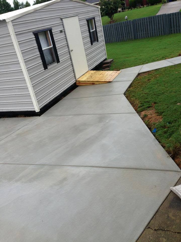 Concrete walkway beside a gray shed, with a small wooden ramp at the door and a fenced yard nearby