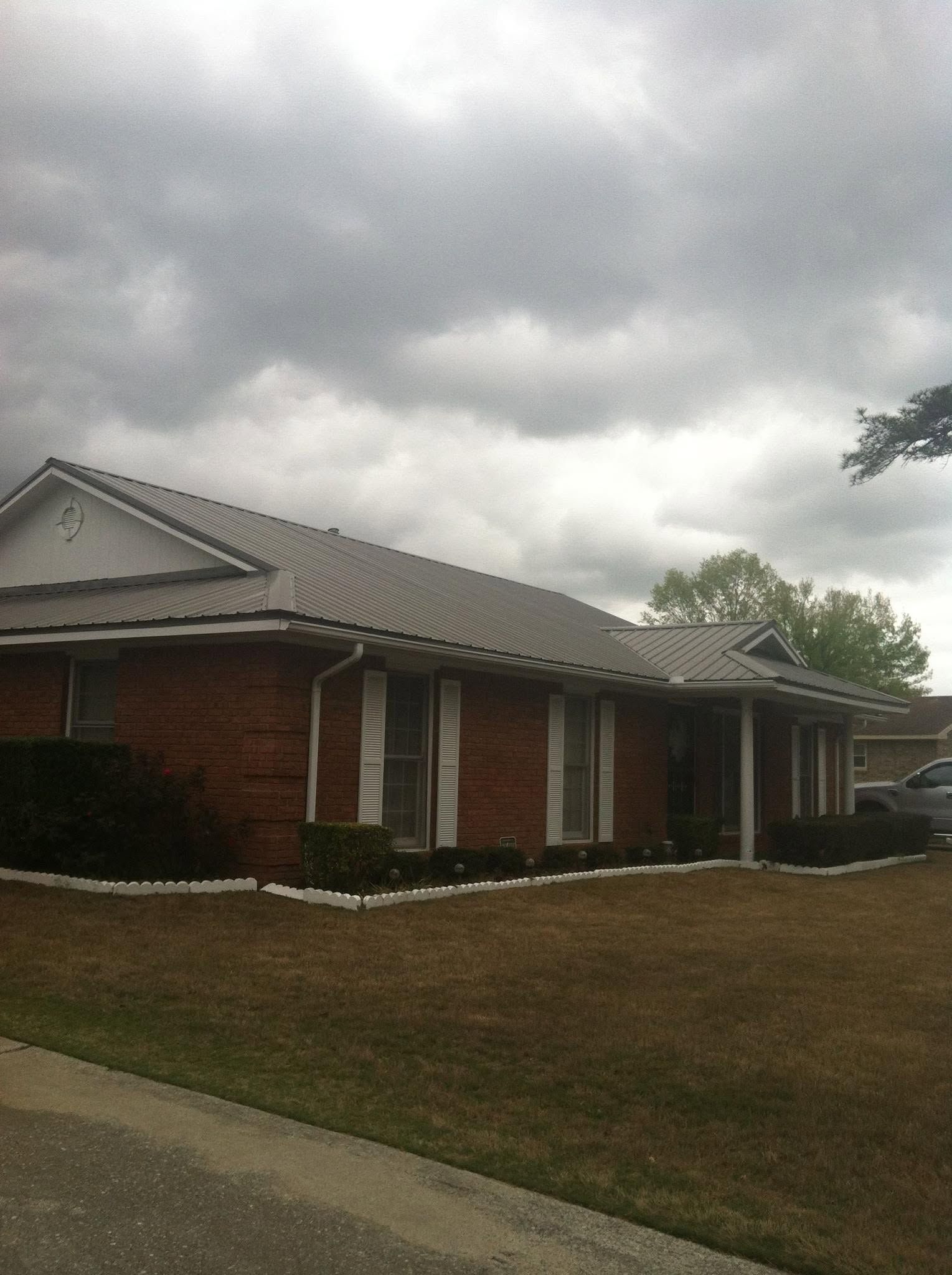 Brick house with a gray roof under cloudy skies, viewed from a grassy lawn by the street