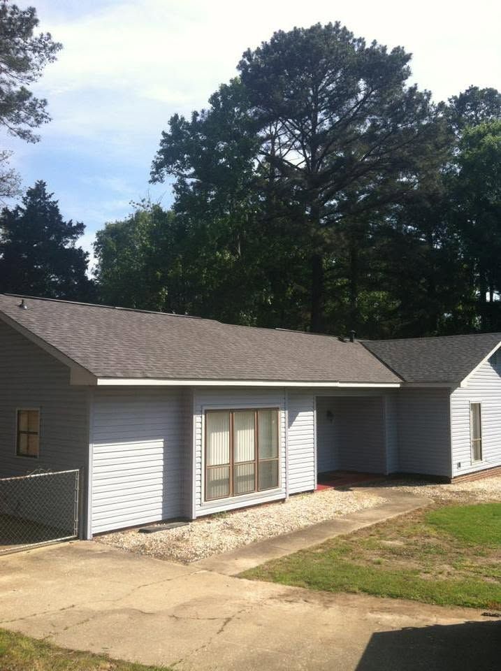 Single-story gray house with white trim, small front window, and fenced yard under tall trees.