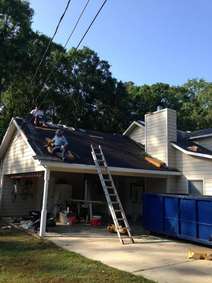 Workers repairing a roof on a house beside a ladder and blue dumpster