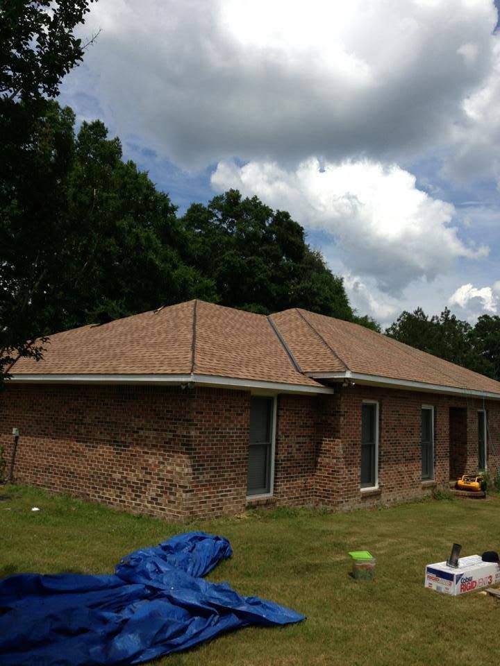 Single-story brick house with tan roof under cloudy sky, with blue tarp and yard items in front.