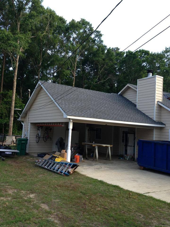Small house with open carport, gray roof, white chimney, blue dumpster, and ladder in the yard.