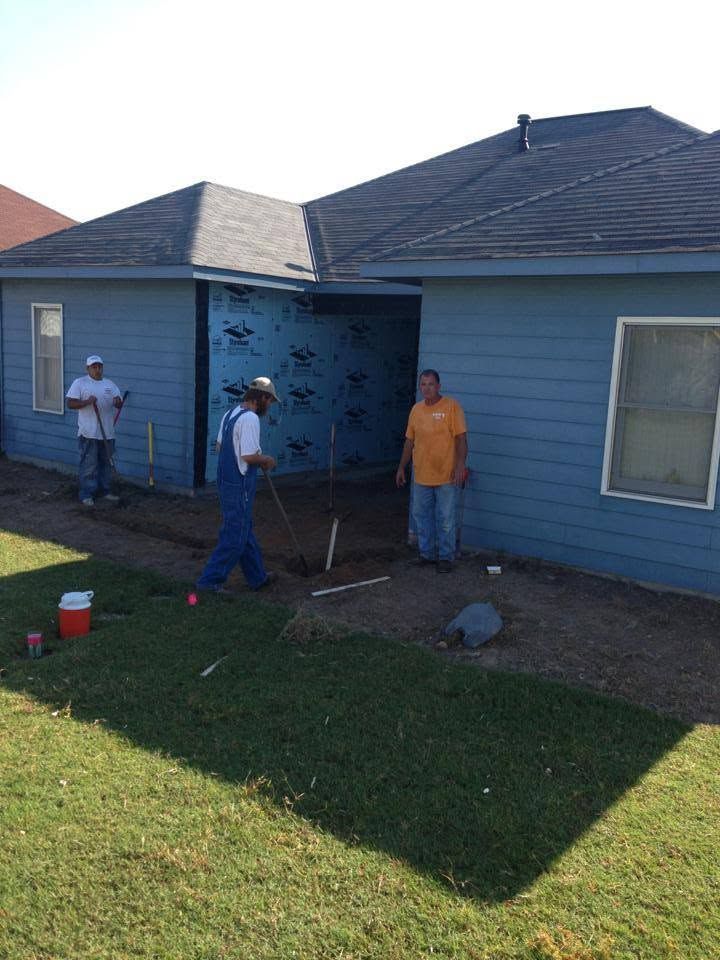Three people standing outside a blue house, one holding a tool near the wall in bright sunlight.