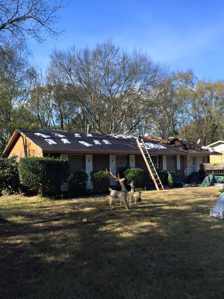 Single-story house with roof damage, ladder, and worker in a yard under a blue sky