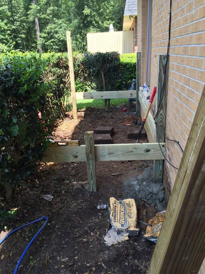 Backyard work area with wooden supports and tools beside a brick house and hedge.