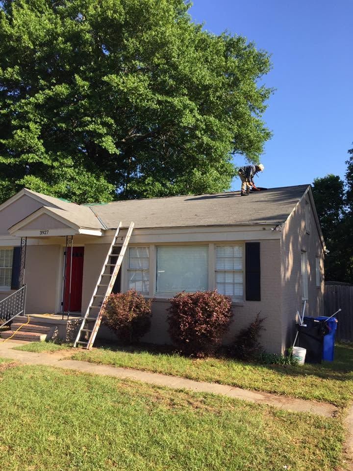 Single-story house with a ladder and a person on the roof near a large tree under a blue sky