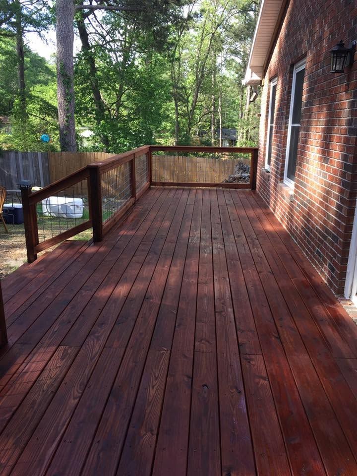 Wooden deck with red-brown boards beside a brick house, bordered by a railing and surrounded by trees