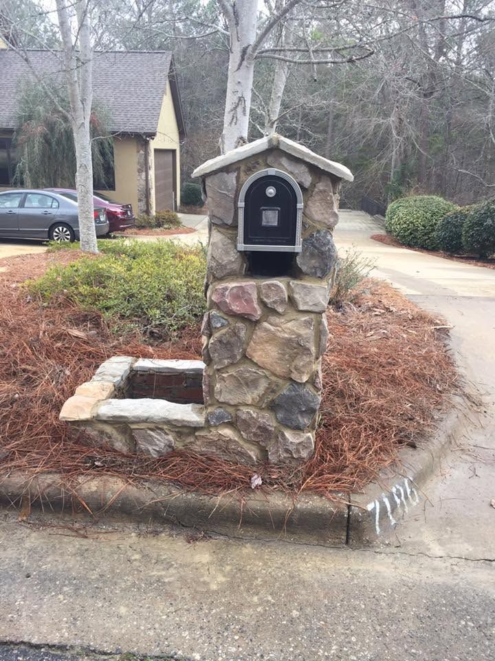 Stone mailbox on a roadside corner with a small planter and a house in the background