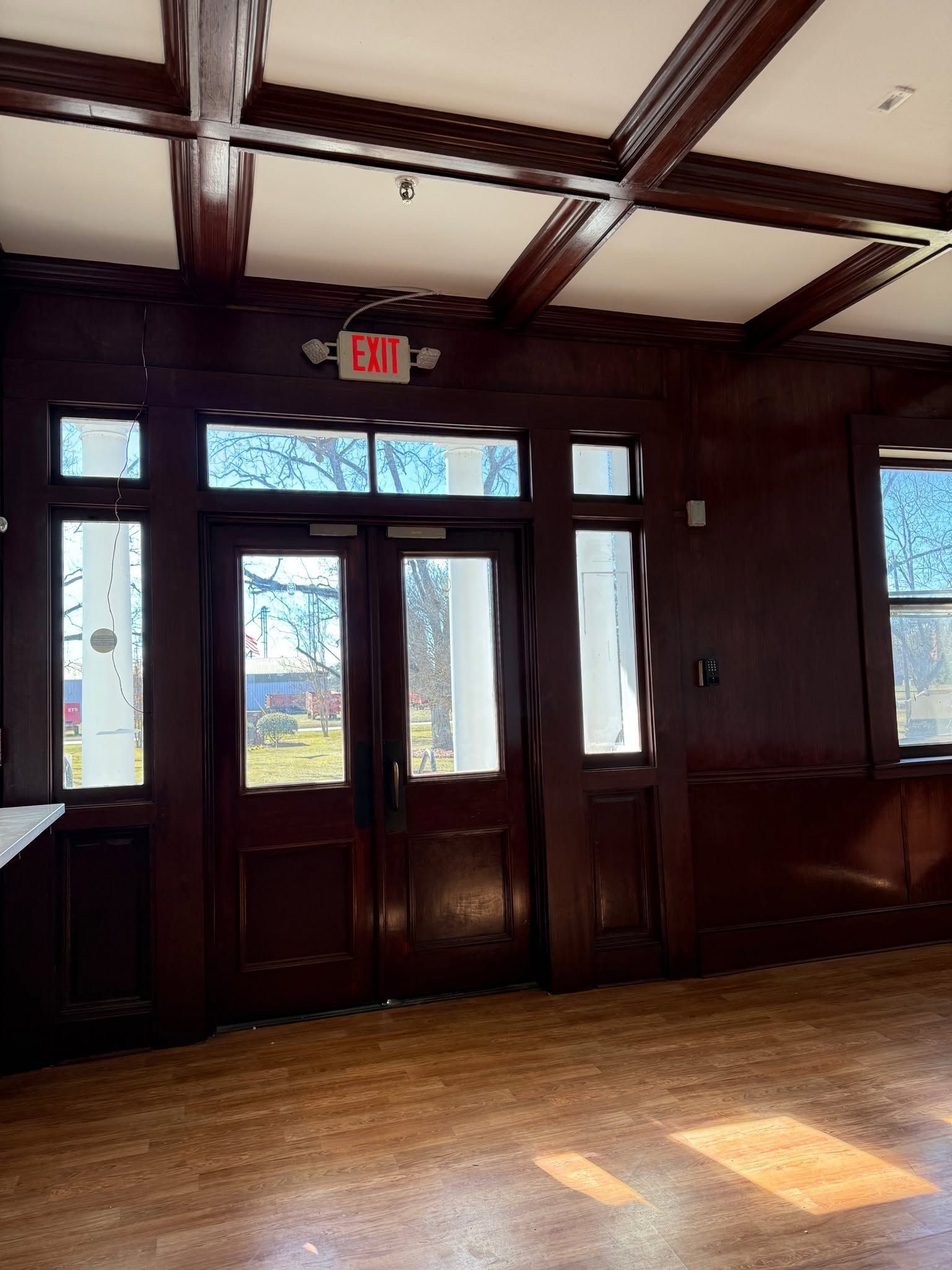 Wood-paneled room with glass double doors and a bright exit sign, sunlight on the floor.