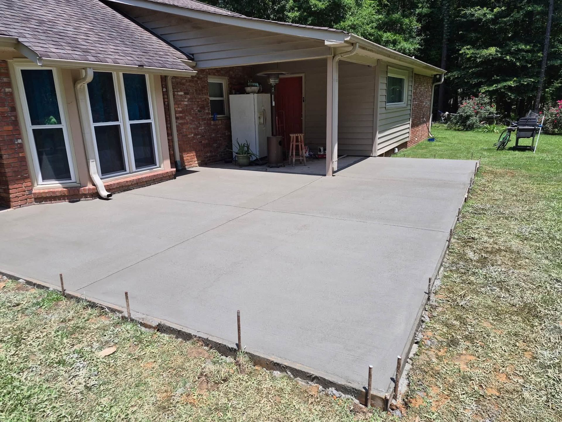 Concrete patio beside a brick house and garage, with a grassy yard and trees in the background.