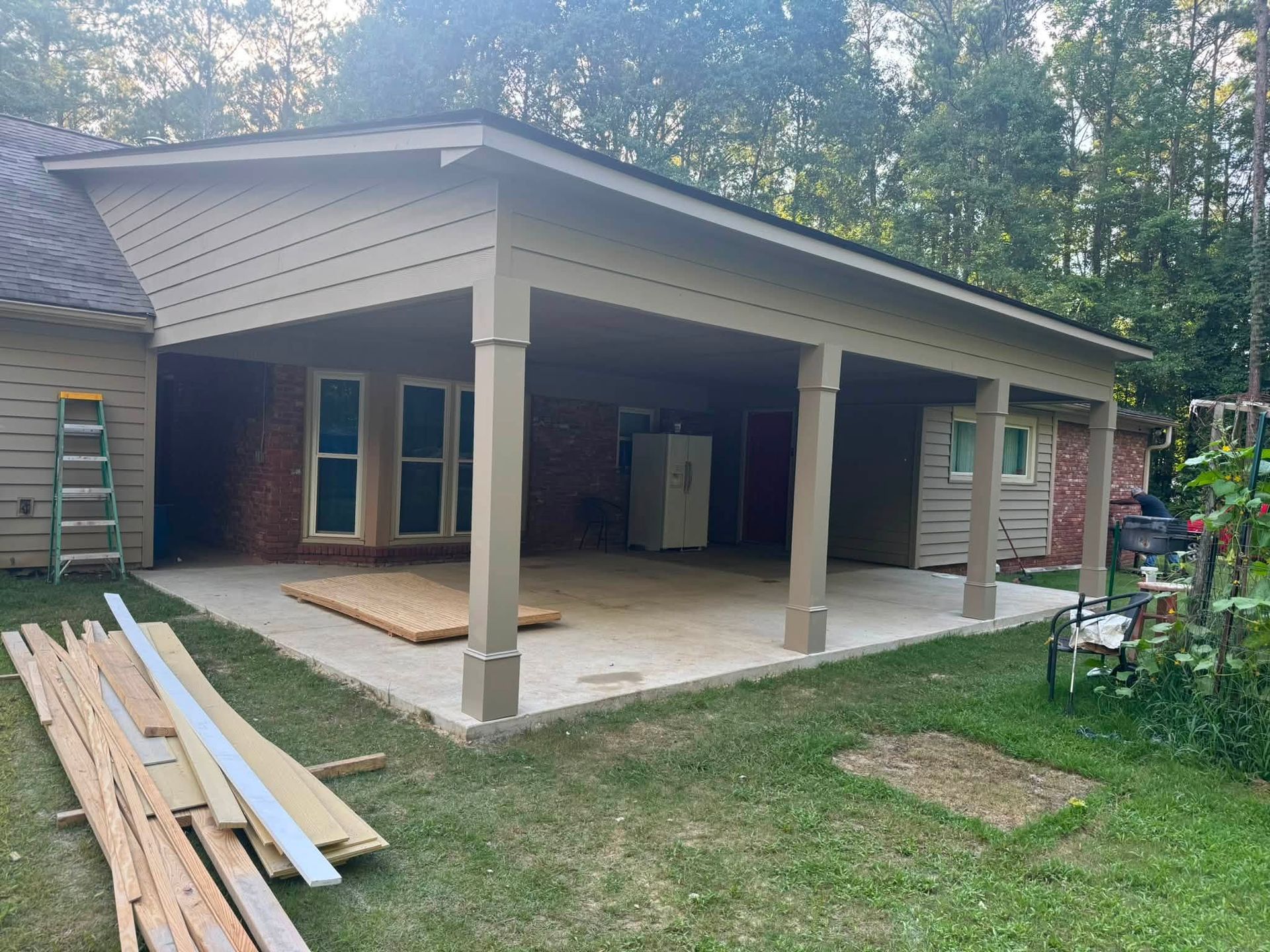 Covered patio construction with white columns and stacked lumber beside a house
