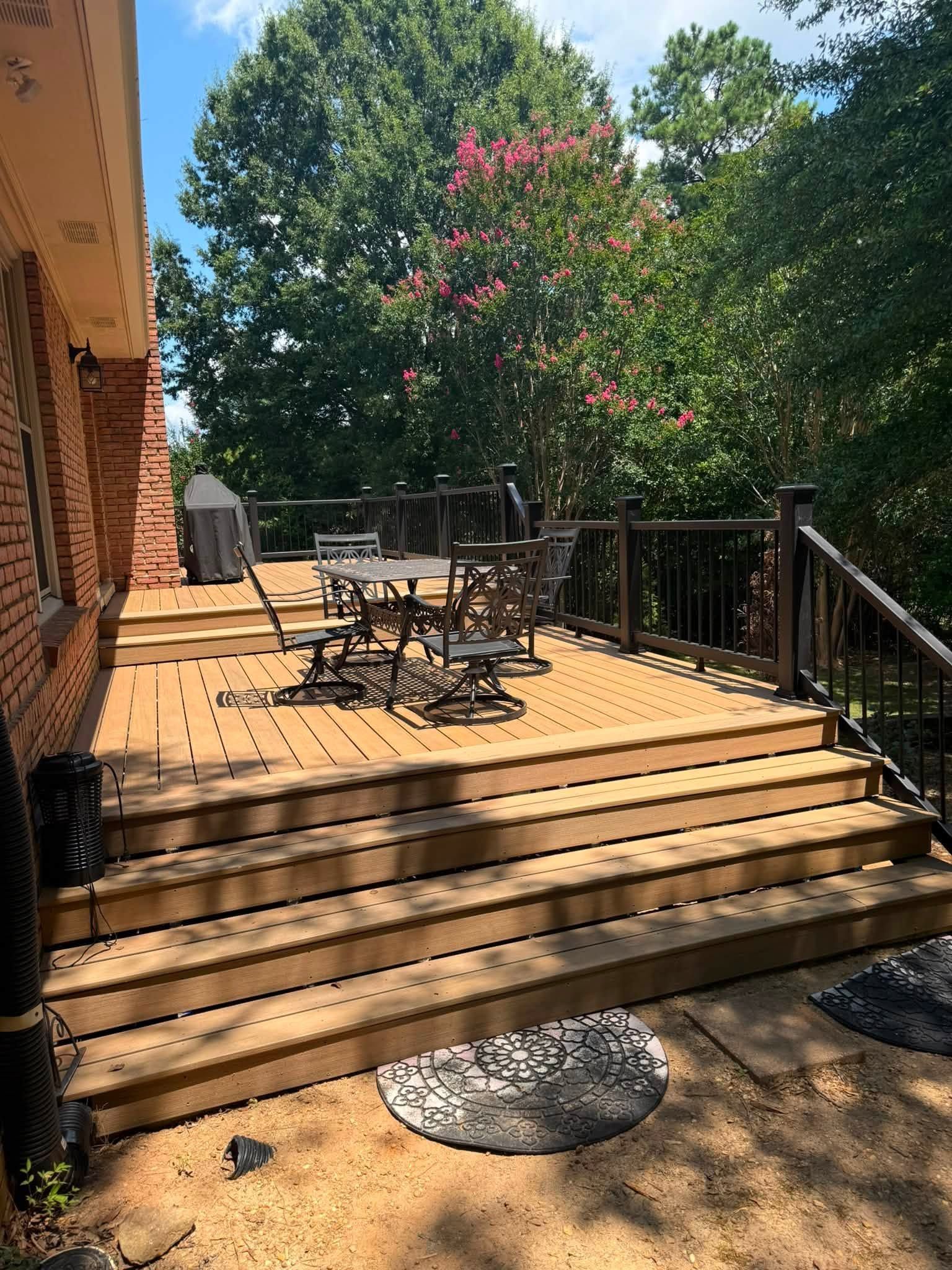 Wooden deck with outdoor table and chairs, steps down to a dirt yard, surrounded by trees and a flowering bush