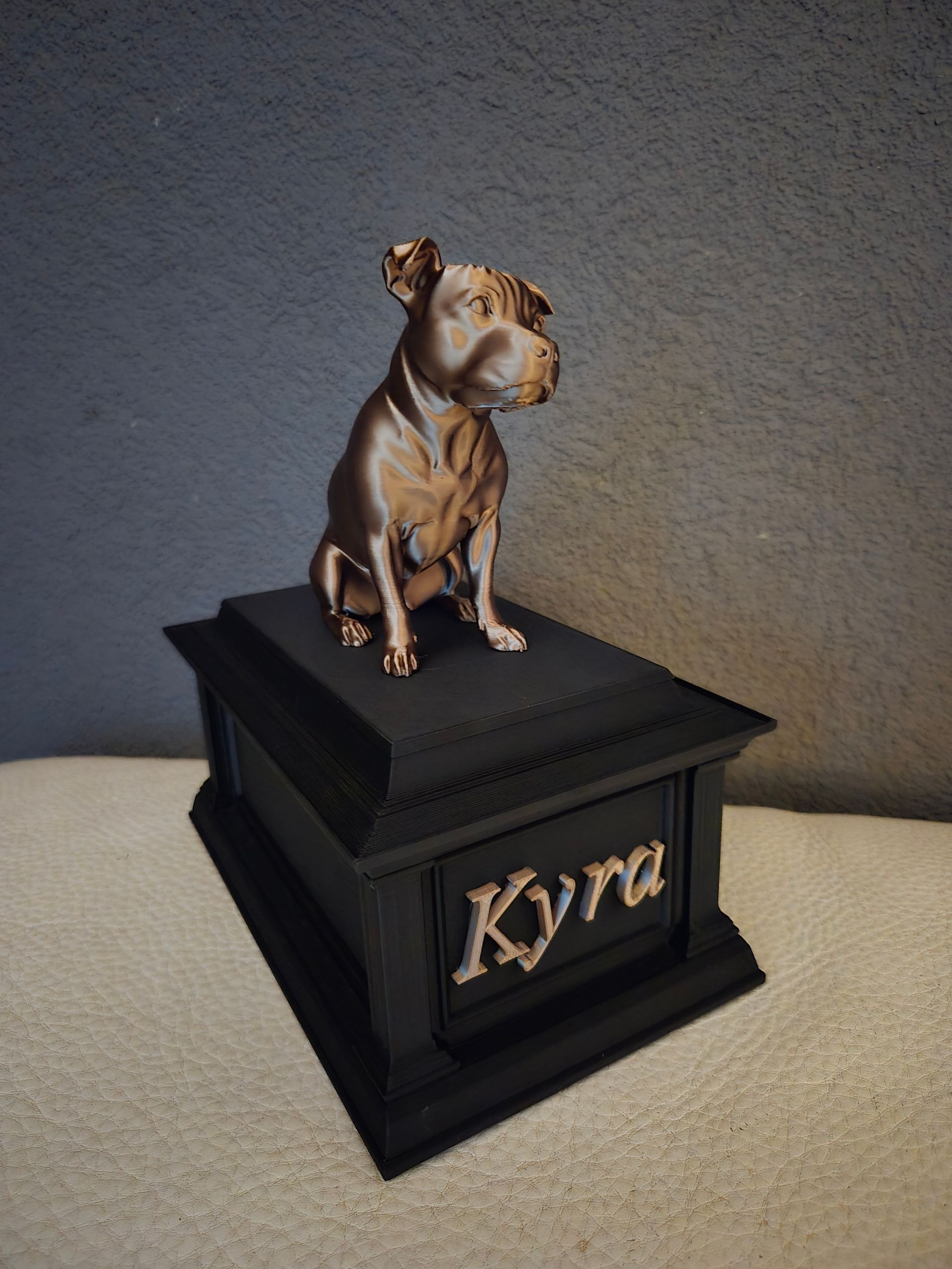 Een metalen, bronskleurig pitbullbeeldje staat bovenop een zwarte gedenkdoos met de naam 