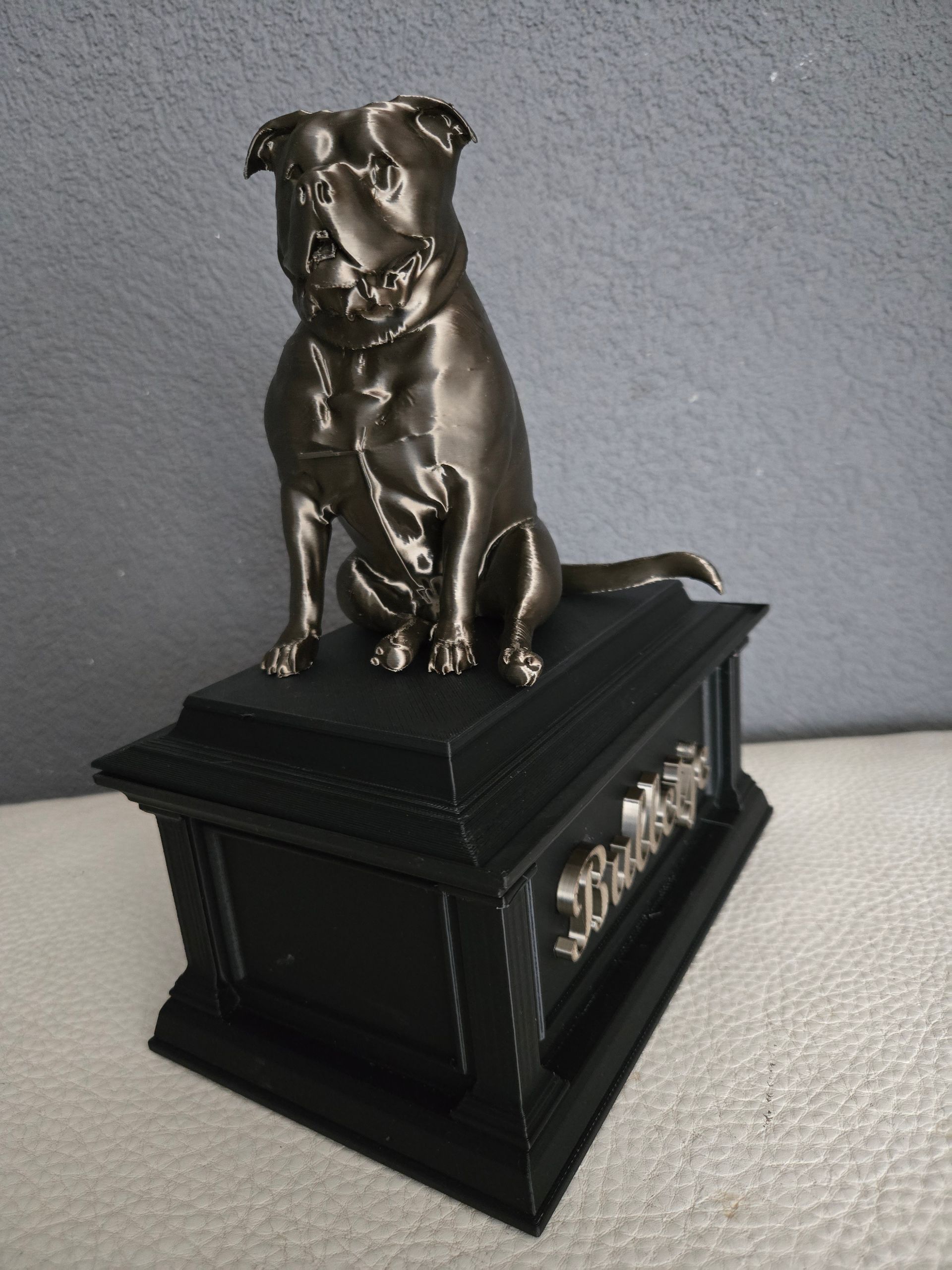 Een metalen, bronzen hondenbeeldje staat bovenop een zwarte, rechthoekige gedenkurn met de naam 