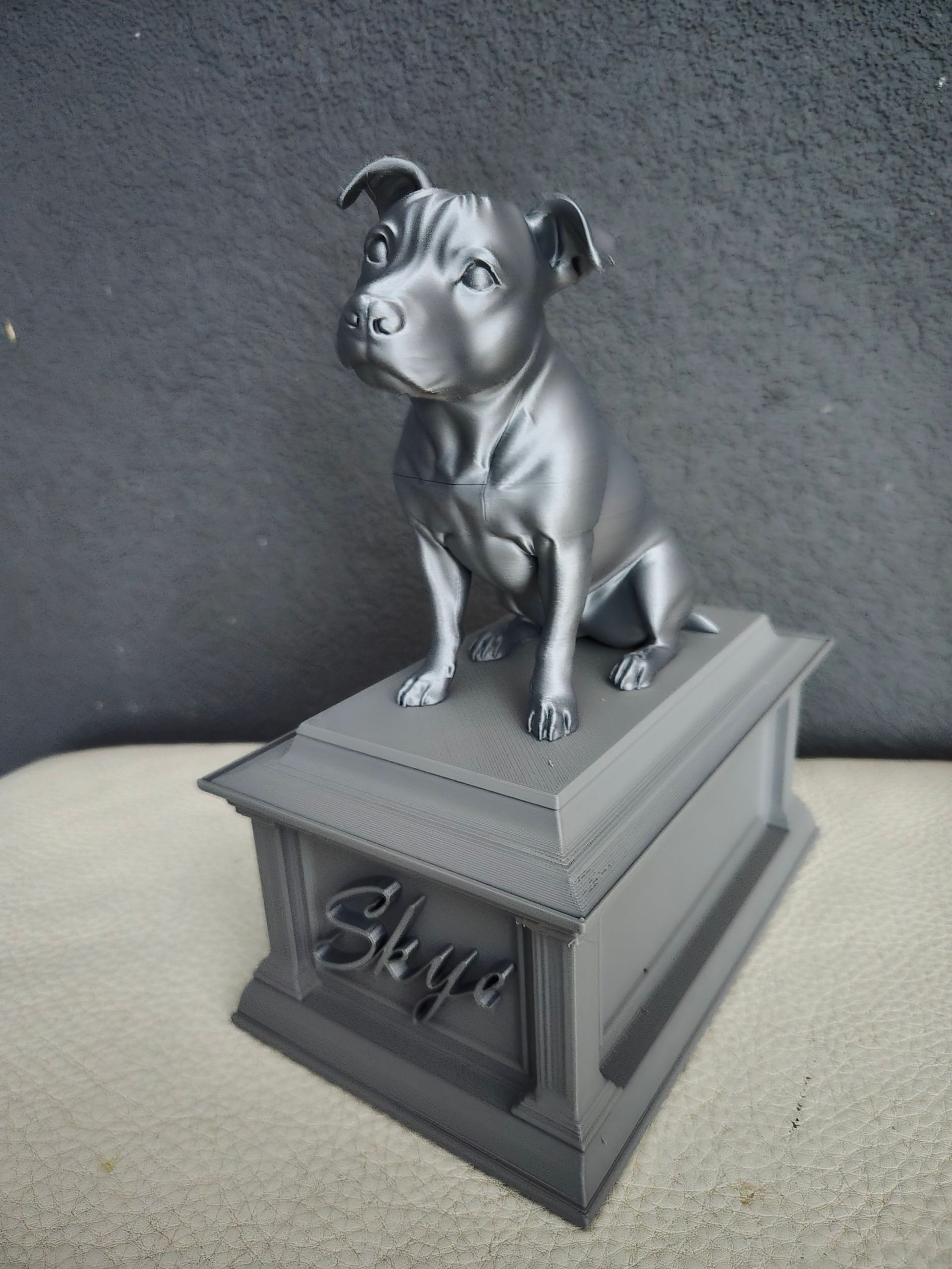 Een grijs, 3D-geprint beeld van een zittende hond, geplaatst op een sokkel met de naam 