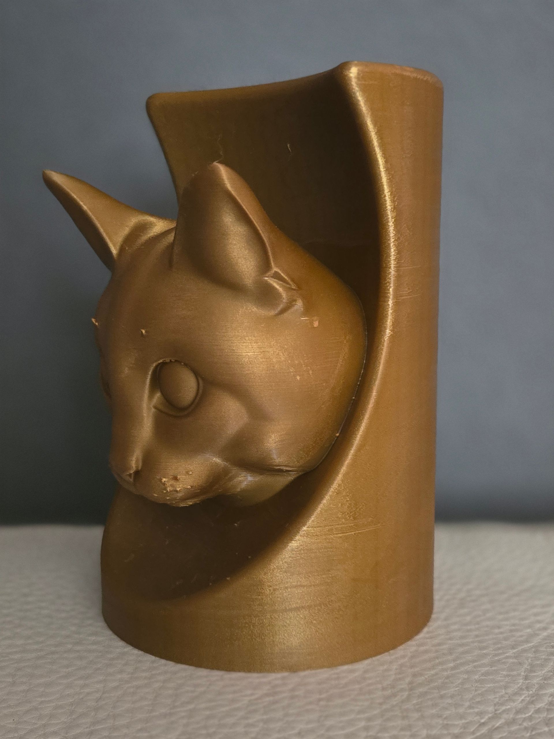 Een goudkleurige, 3D-geprinte cilindervormige container met een gebeeldhouwde kattenkop die uit de voorkant steekt.