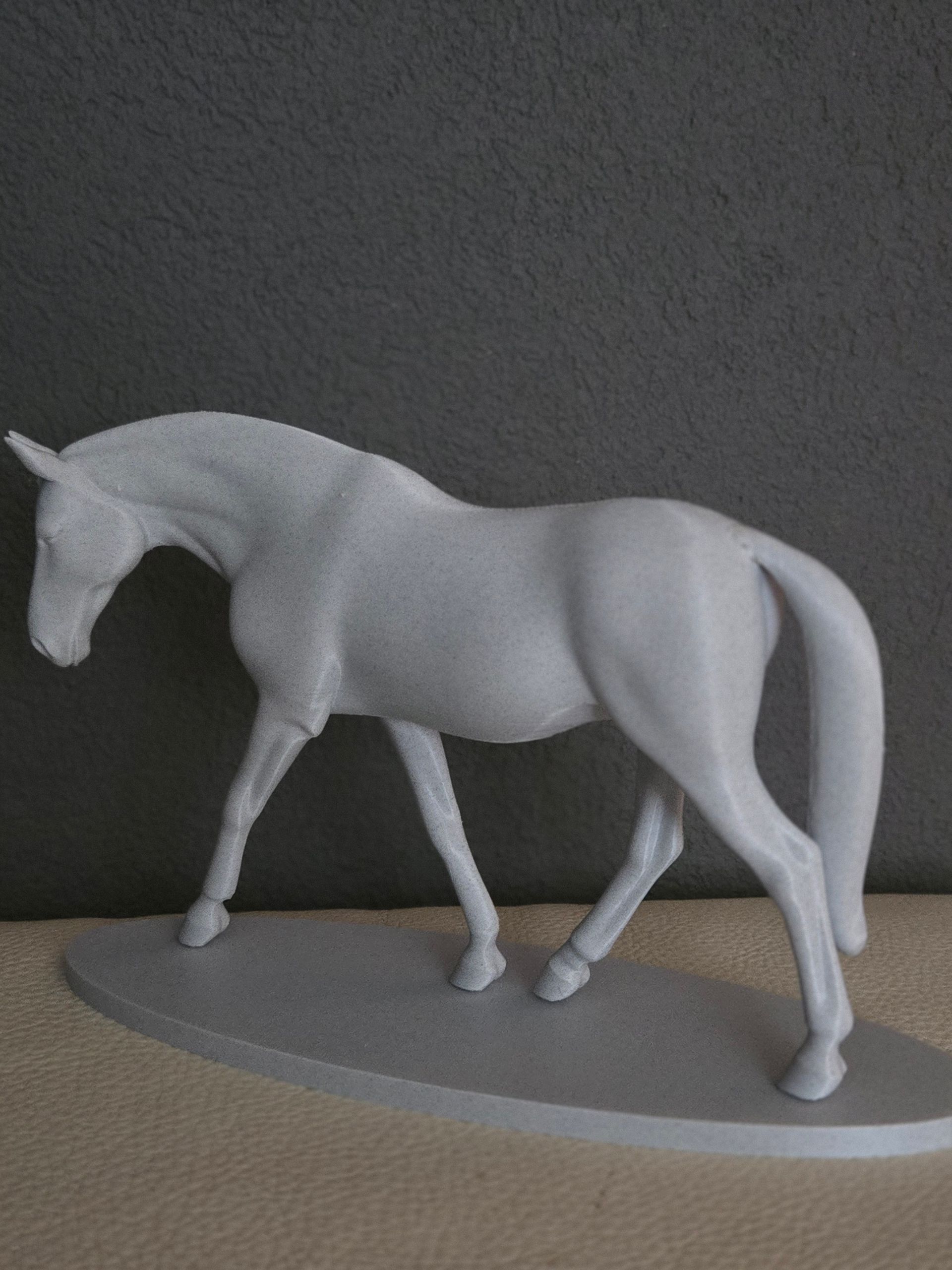Een eenvoudig grijs beeldje van een paard dat op een ovale sokkel staat tegen een neutrale achtergrond.