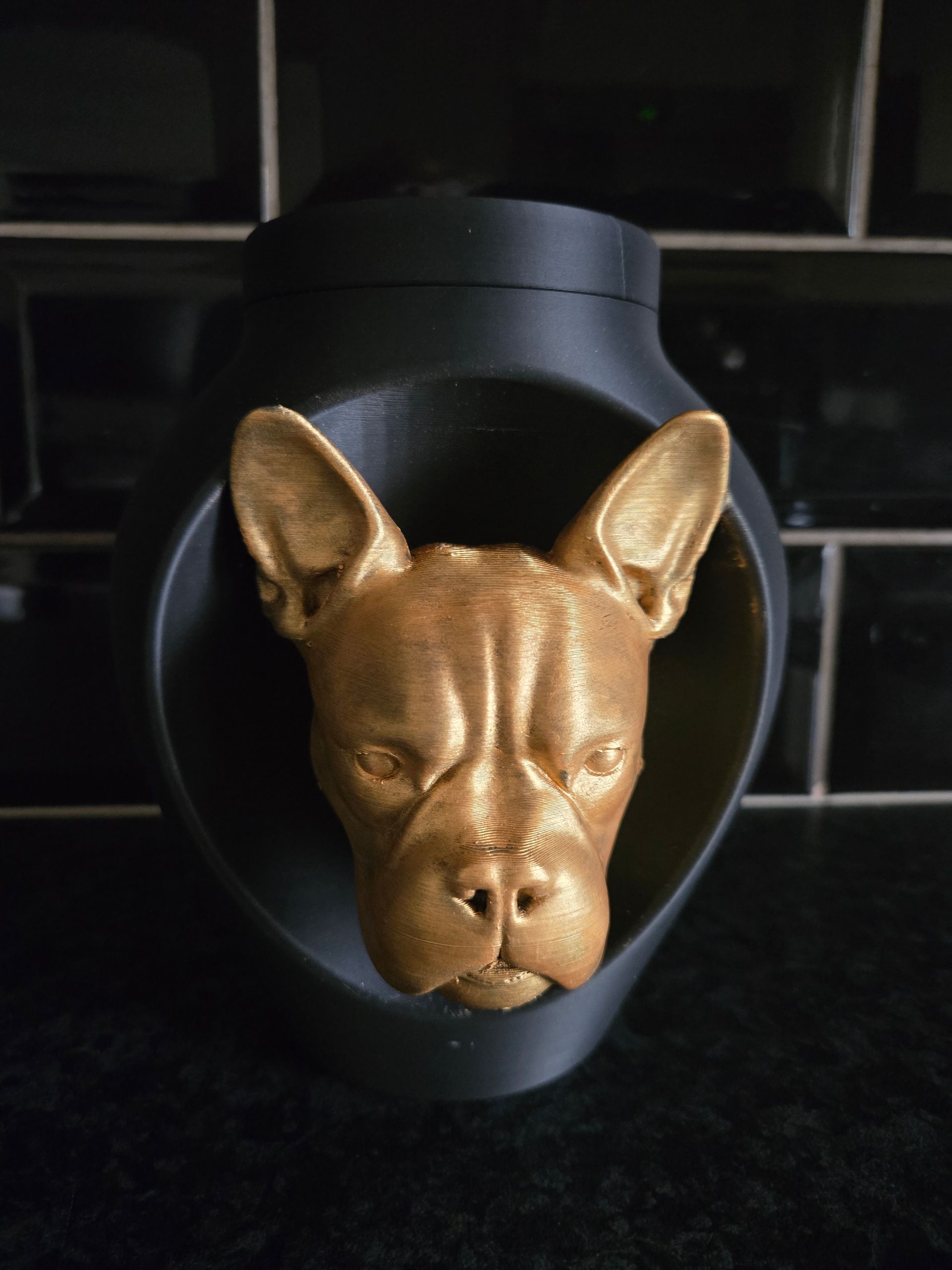 Een goudkleurig, 3D-geprint beeldje van een Franse bulldogkop, gemonteerd op een matzwarte, ronde sokkel.
