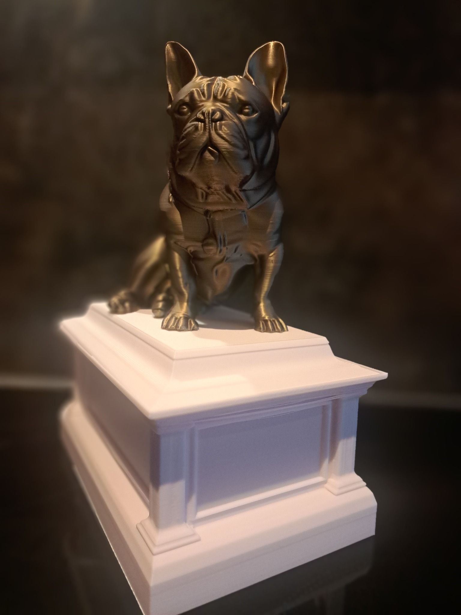 Een bronskleurig beeldje van een Franse bulldog staat op een rechthoekig, gelaagd wit voetstuk.